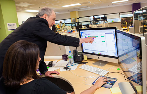 Woman setting before Mac screens, man reaching in front of her to point at something on screen